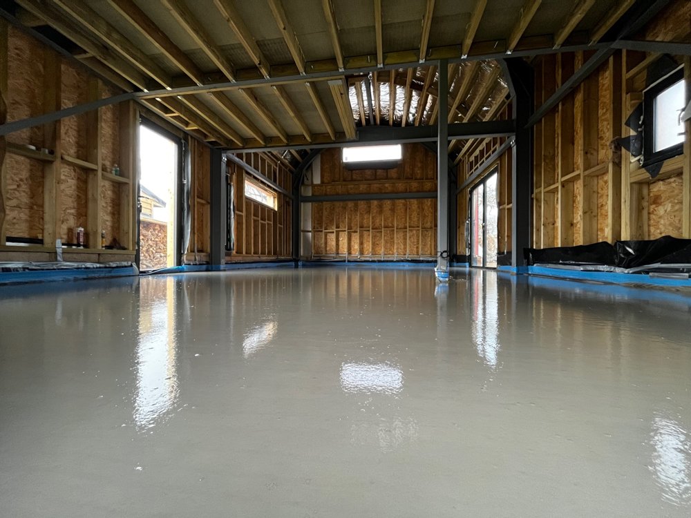 Indoor construction site with a fresh concrete floor, exposed wooden framing, and windows, showing the structure of a building under construction.