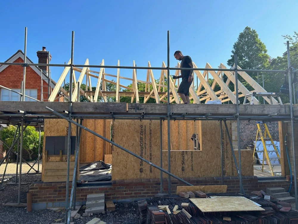 A man is working on the wooden frame of a house under construction, standing on scaffolding outside during daytime with a house and trees in the background.