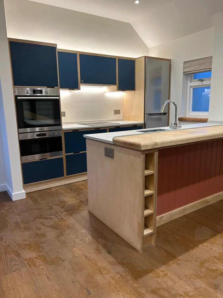 Modern kitchen with blue upper cabinets, light wood lower cabinets, a white countertop, built-in stainless steel oven, and a refrigerator. Wooden flooring and a window with a blind.