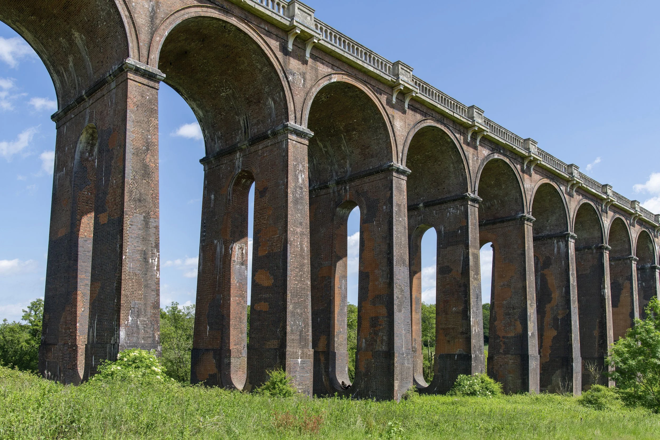 A large brick bridge with multiple arches spanning over a green area with trees and bushes, under a blue sky with some clouds.
