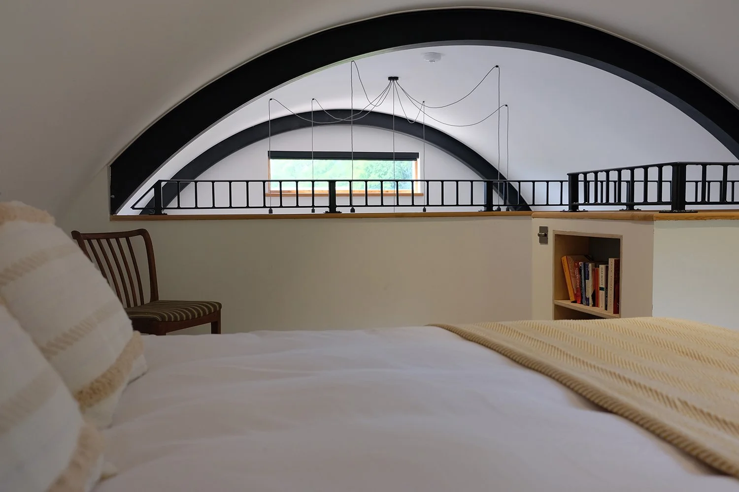 A cozy bedroom with a large, curved ceiling, a bed with white linens and beige striped blankets, an open bookshelf, a wooden chair, a small window with green trees outside, and modern lighting fixtures hanging from the ceiling.