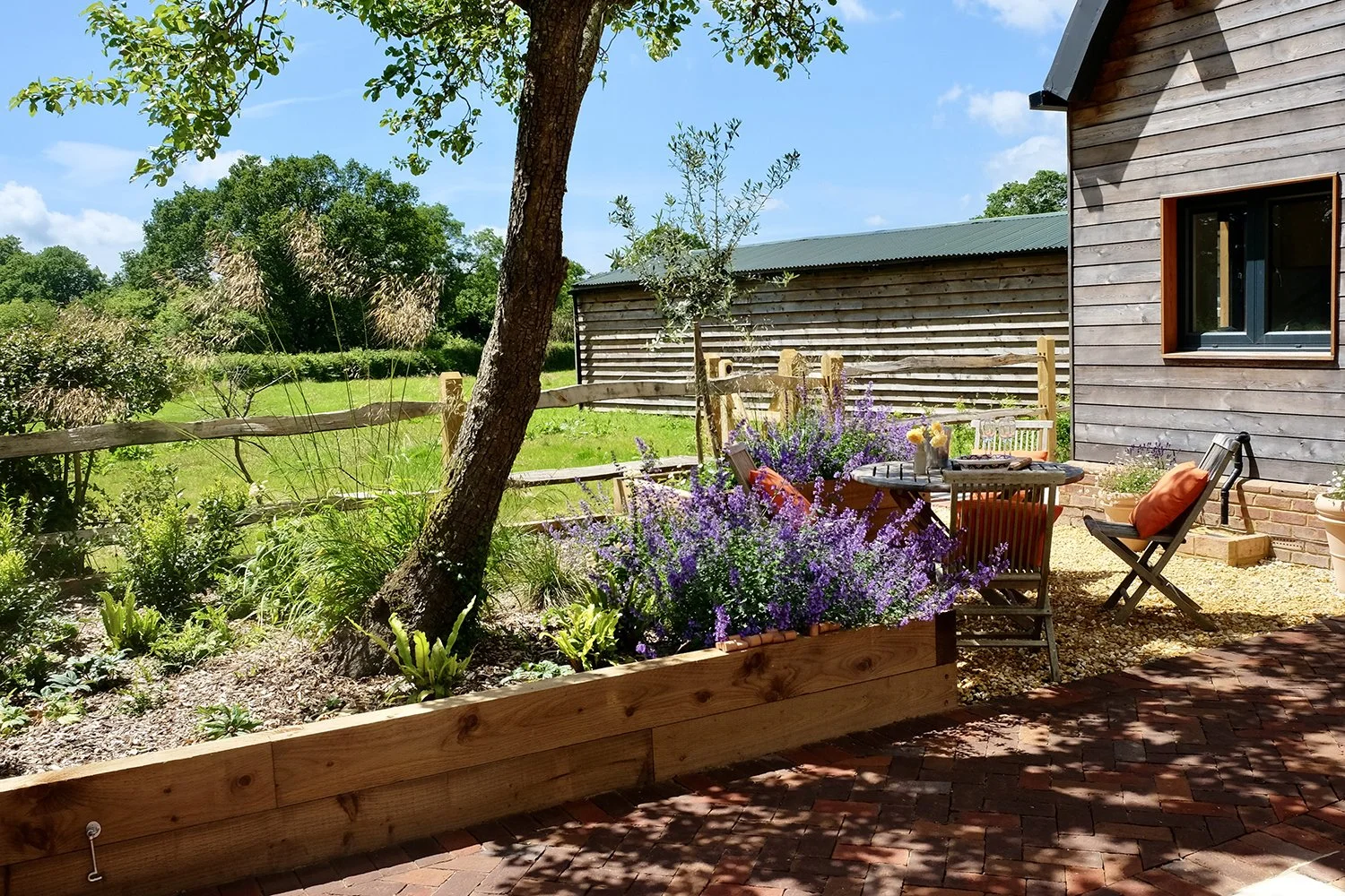 A cozy garden patio with a round table, chairs with cushions, potted plants, purple flowers, trees, a wooden fence, and a wooden house wall in the background, under a blue sky with clouds.