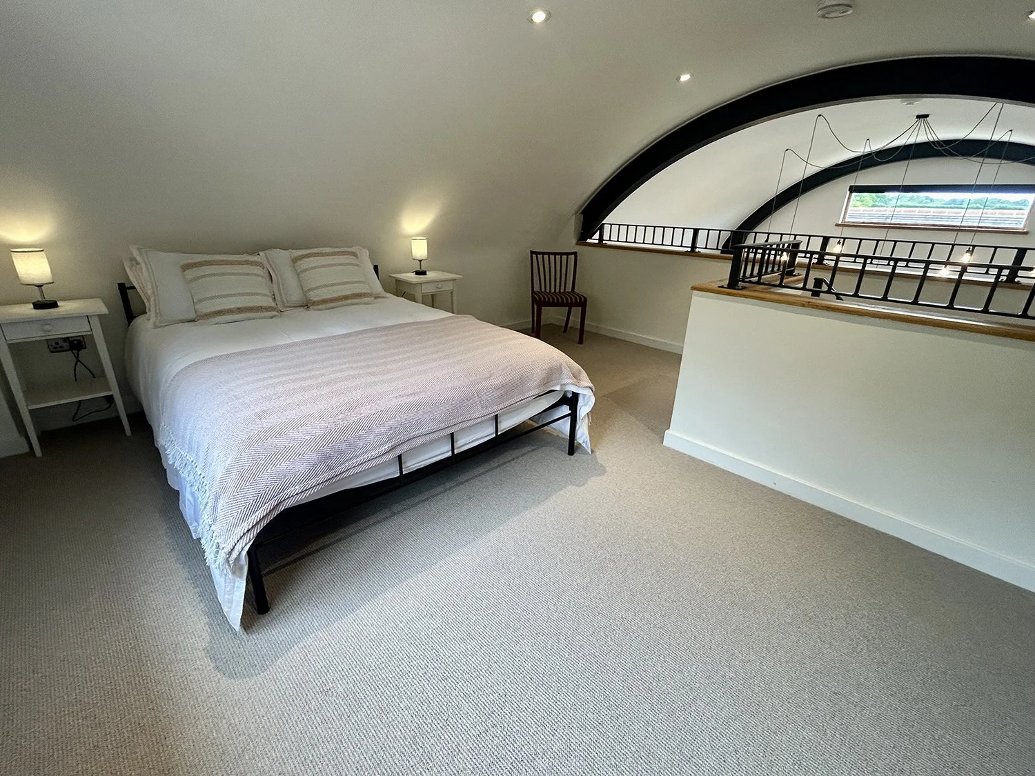 A bedroom with a sloped ceiling, a white bed with striped pillows, two white nightstands with lamps, a dark wooden chair, beige carpet, and a loft railing with a window in the background.