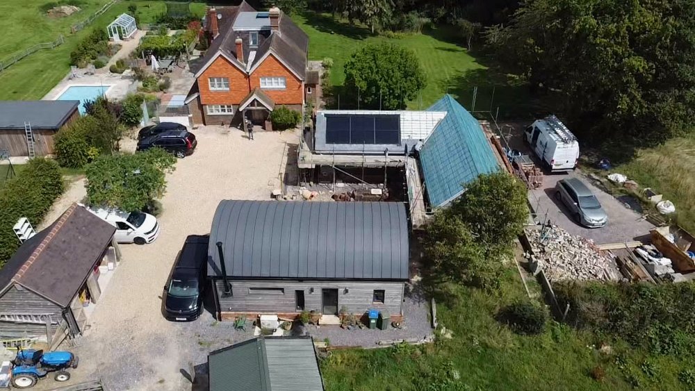 Aerial view of a residential backyard with a main house, a greenhouse under construction, a shed, multiple parked cars, a small swimming pool, and various gardening and construction equipment.