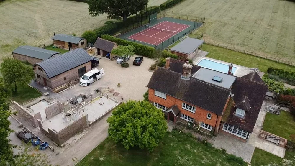 Aerial view of a large property with a brick house, a swimming pool, tennis court, small outbuildings, a detached garage, a gravel driveway, and surrounding green fields.