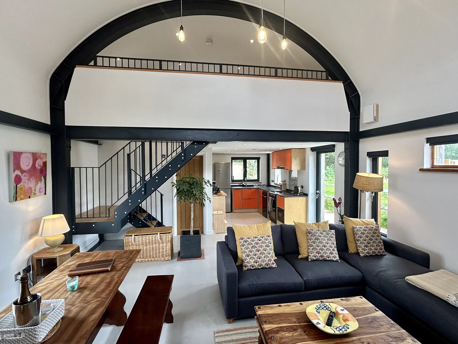 Interior of a modern, open-plan living space with black metal beams and a loft area, a dark gray sectional sofa with yellow and patterned pillows, a wooden coffee table, a dining table with a partner chair and stool, a kitchen with orange and beige c
