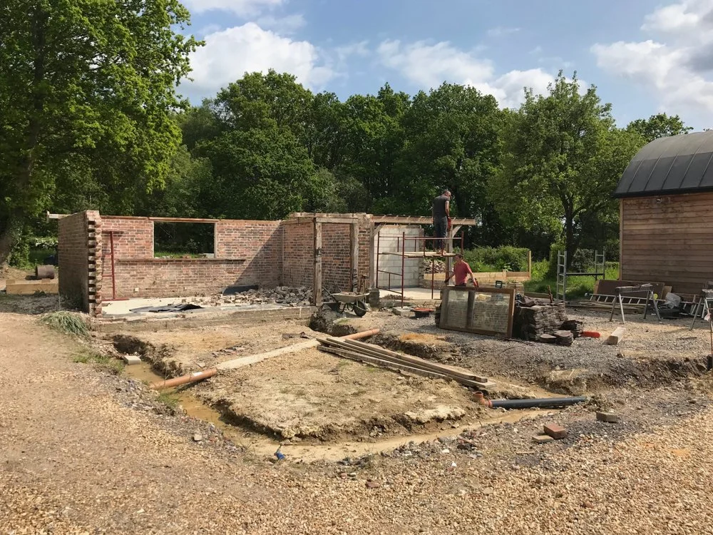 Construction site with partially built brick walls and workers installing a wooden roof, surrounded by trees and construction materials.