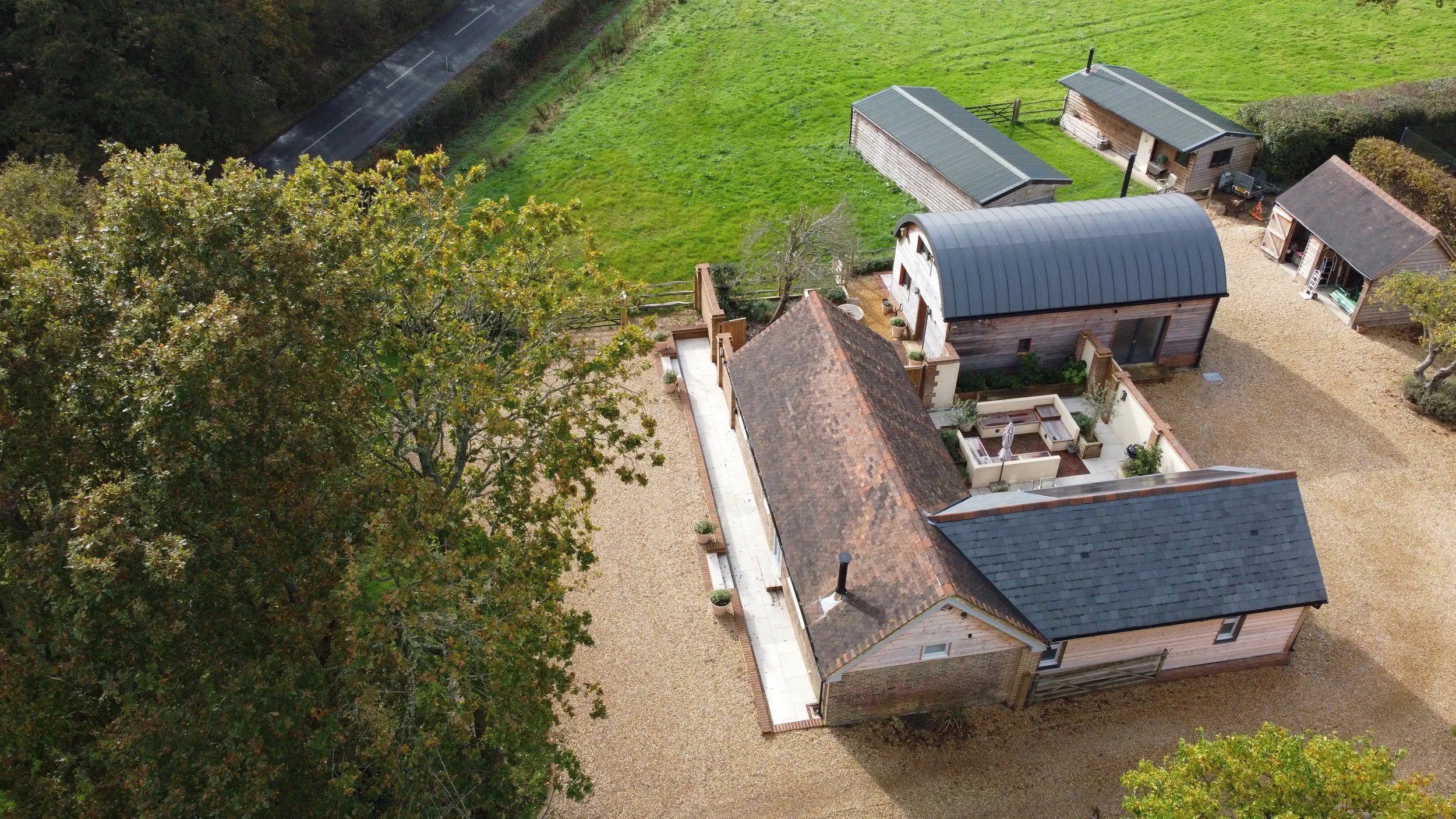 Aerial view of a residential backyard showing a gravel driveway, a patio with outdoor furniture, a large tree, and a small green lawn area with small outbuildings or sheds.
