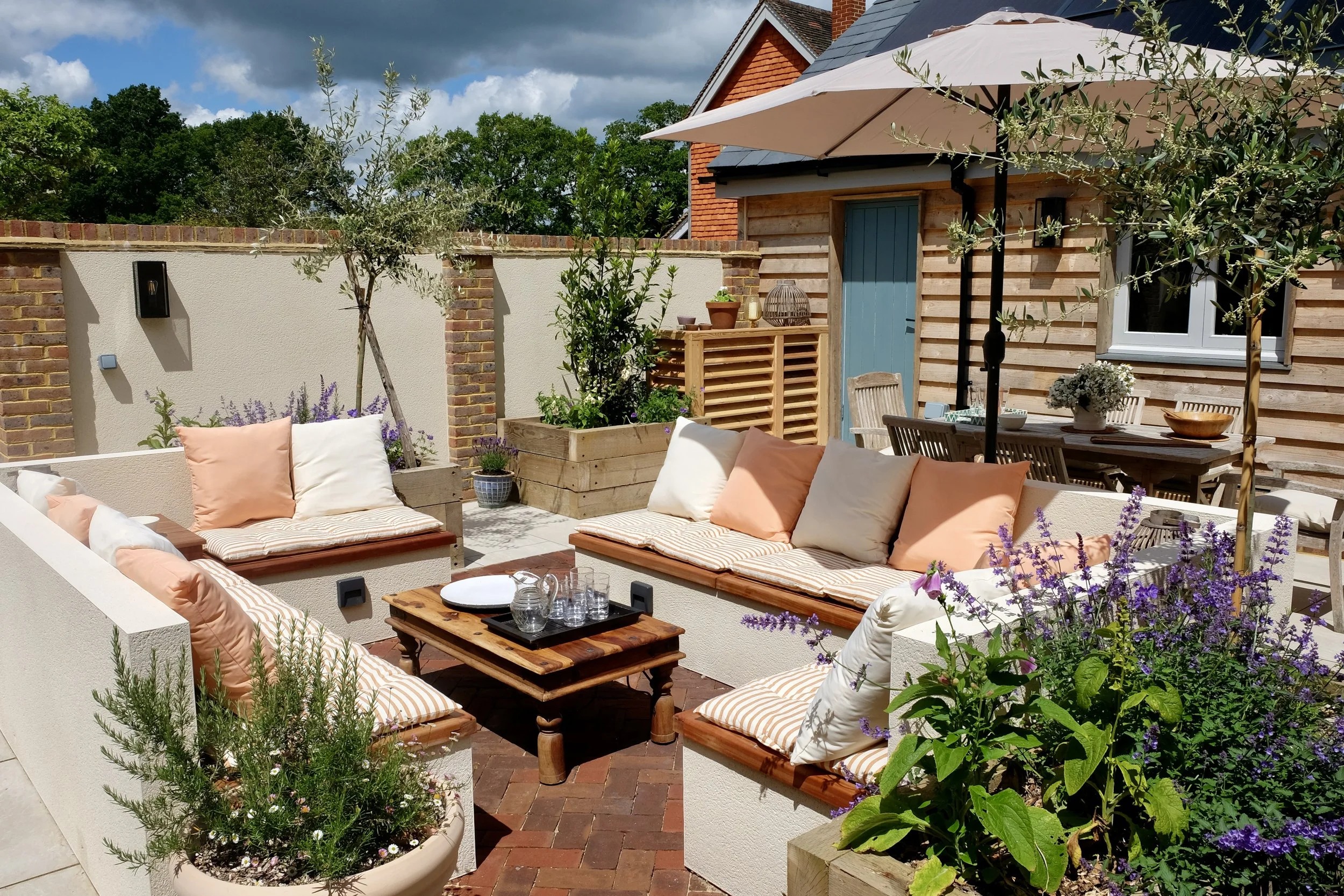 An outdoor patio area featuring cushioned seating with pink and white pillows, a wooden coffee table with drinkware, potted plants, a large umbrella, and a wooden shed with a blue door. The space is enclosed by a brick and stucco wall, with trees and a house with a sloped roof in the background.