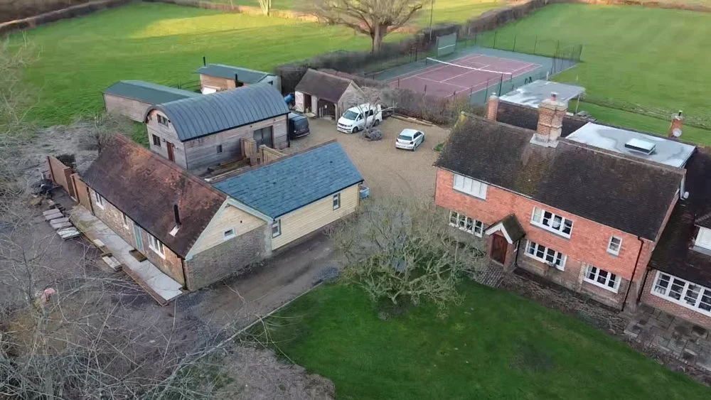 Aerial view of a residential property with a main brick house, a small yellow outbuilding, a curved-roof shed, tennis courts, and surrounding garden and lawn area.