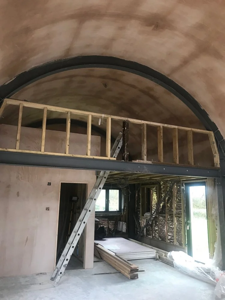 Interior of a building under construction with a curved ceiling, an upstairs wooden railing, ladder, and construction materials inside.