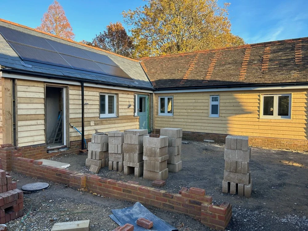 Backyard construction site with stacks of concrete blocks, a partially built brick wall, and a house with solar panels installed on the roof. The house has yellow siding and several windows, with a backdrop of trees and a blue sky.