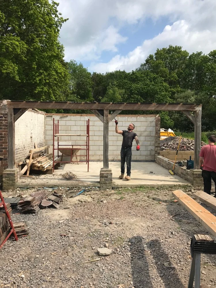 Construction site with two men working, one standing and pointing upward, the other with his back turned, amidst building materials and partially constructed walls under a wooden framework, surrounded by trees and a partly cloudy sky.