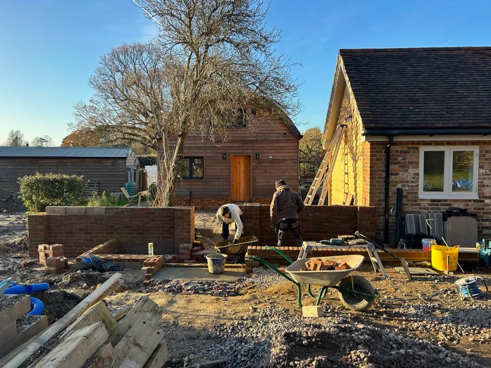 Two people are constructing a brick wall in a backyard during the daytime, with a finished house and a wooden shed visible in the background.