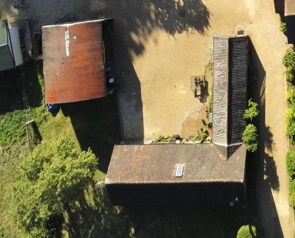 An aerial view of a backyard with a brick house, a metal-roof extension, a gravel area with some garden plants, and trees casting shadows.