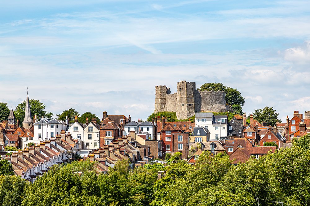 A view of a town with many houses and a large historic castle on a hill in the background, under a partly cloudy sky.