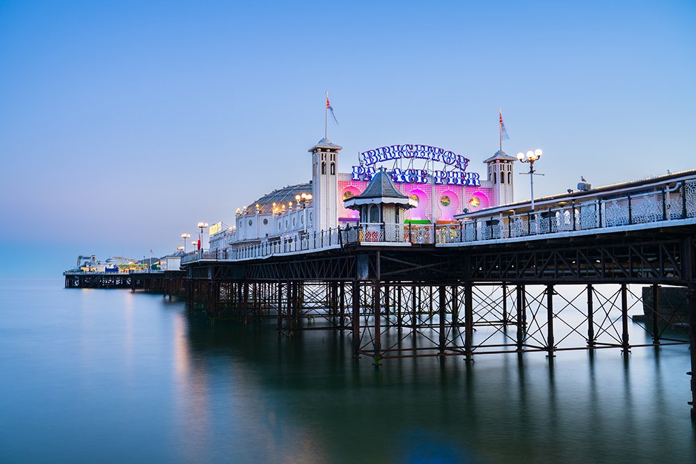 Brighton Pier extending over calm water under a clear sky, with colorful signage and lights along the promenade.