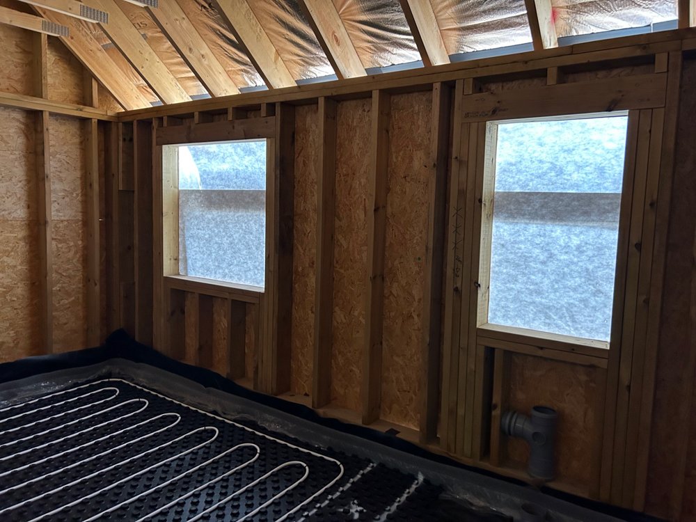 Interior of a building under construction with wooden framing, two windows covered with protective plastic, and a radiant floor heating system installed on the floor.