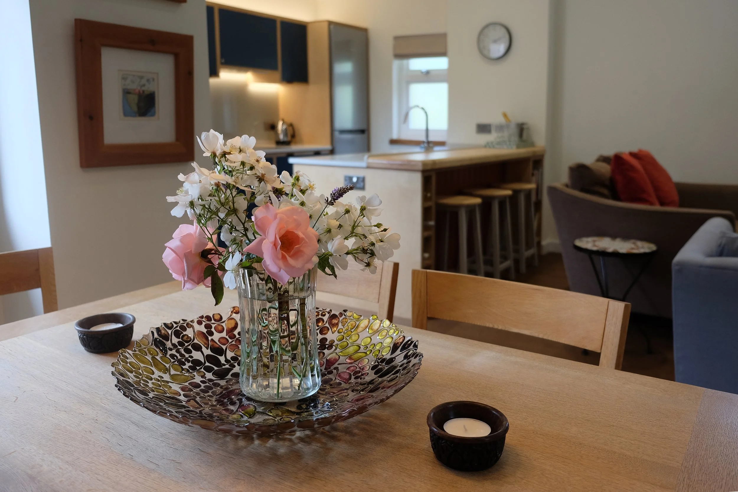 A kitchen with a wooden dining table, a floral centerpiece in a glass vase on a decorative plate, two black candle holders, a kitchen counter with stools, a window, a wall clock, and various kitchen appliances.