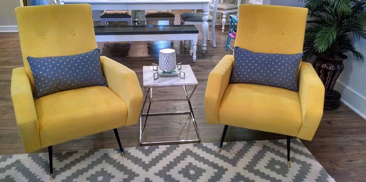Two yellow armchairs with gray polka dot pillows facing each other with a small white marble side table in between, on a gray and white patterned rug in a living room.