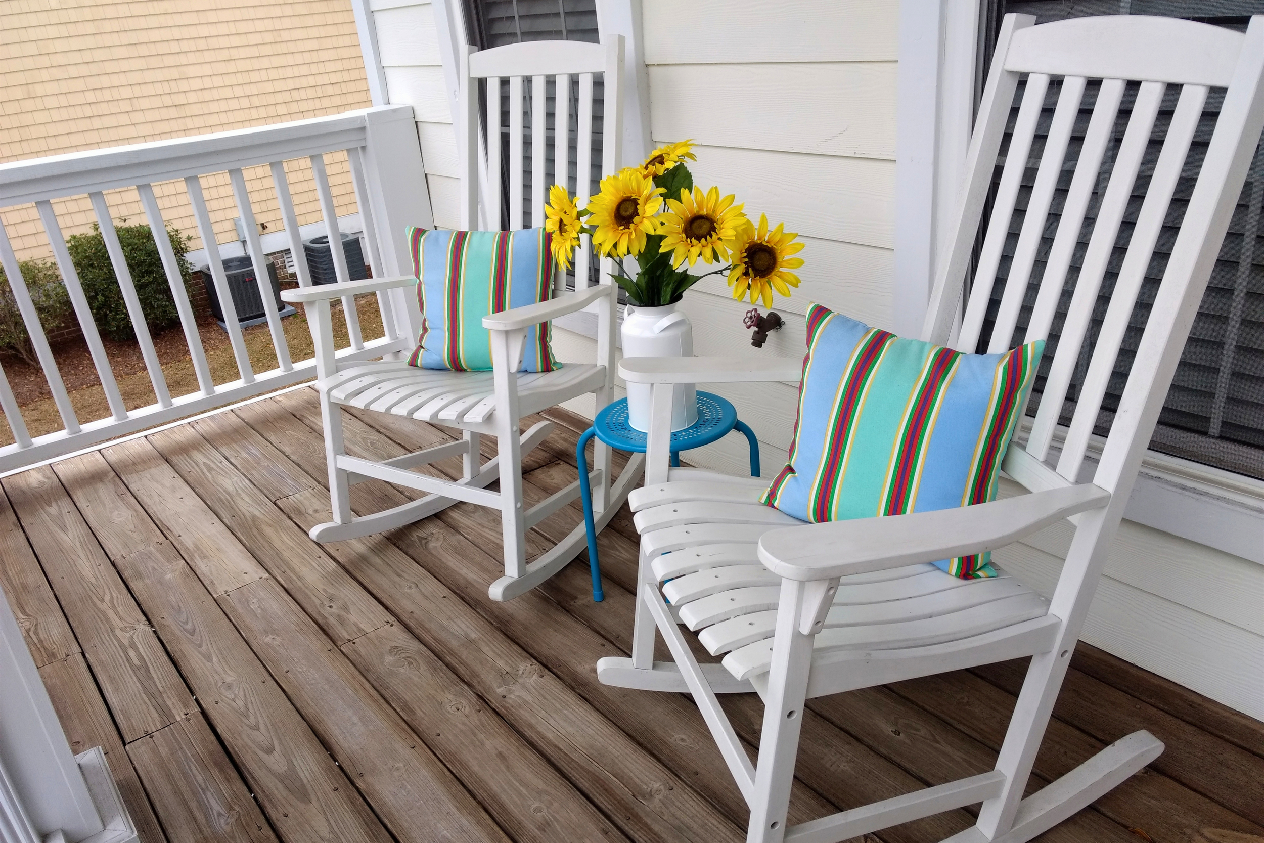 A small porch with wooden flooring, three white rocking chairs with colorful striped pillows, a white side table with a pitcher holding bright yellow sunflowers, a hose faucet on the wall, and a background of beige and white exterior walls.