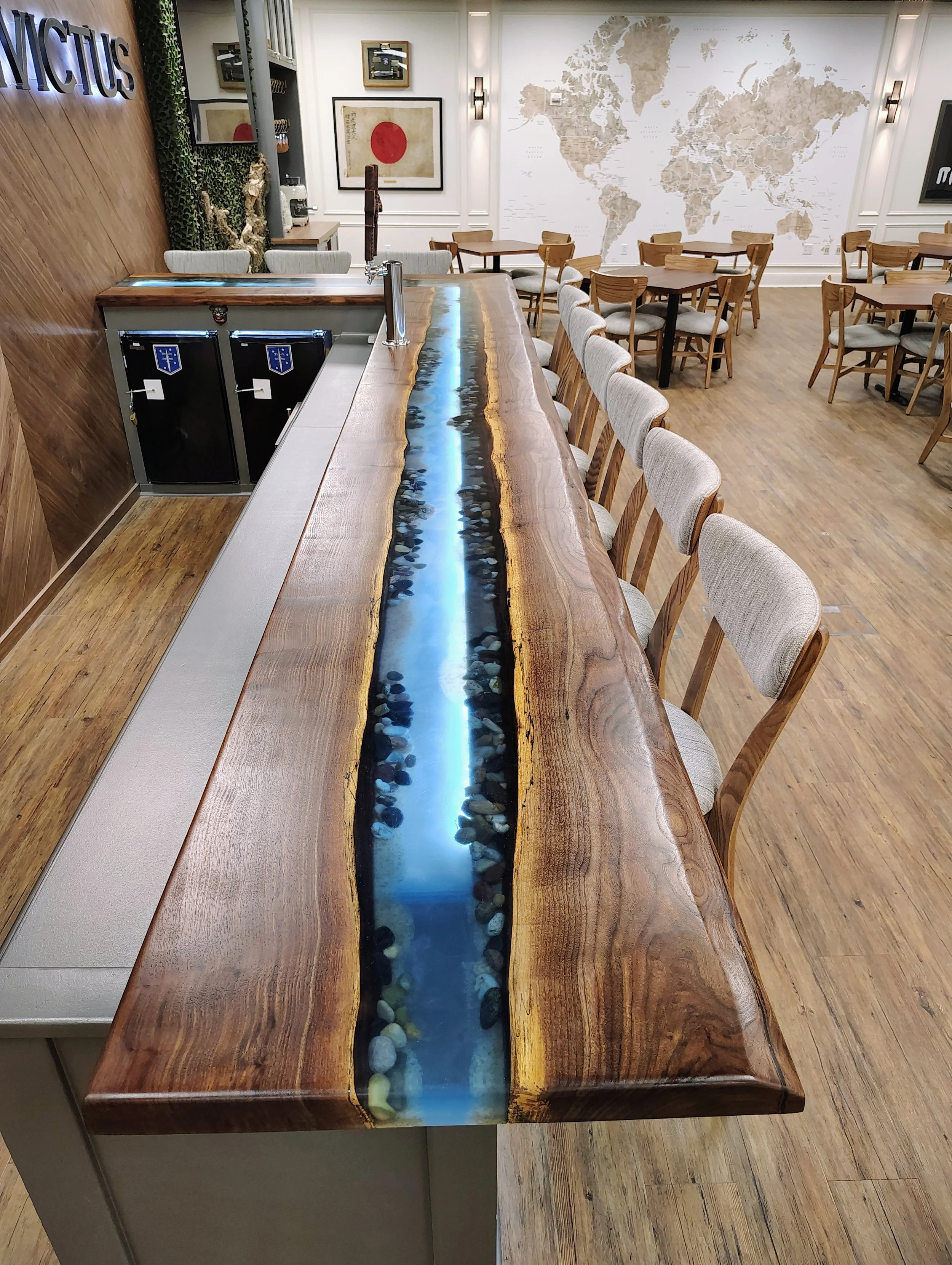 A wooden bar with a river-like glass inlay running through the center, rocks inside the glass, in a modern restaurant or cafe with wooden flooring and tables.