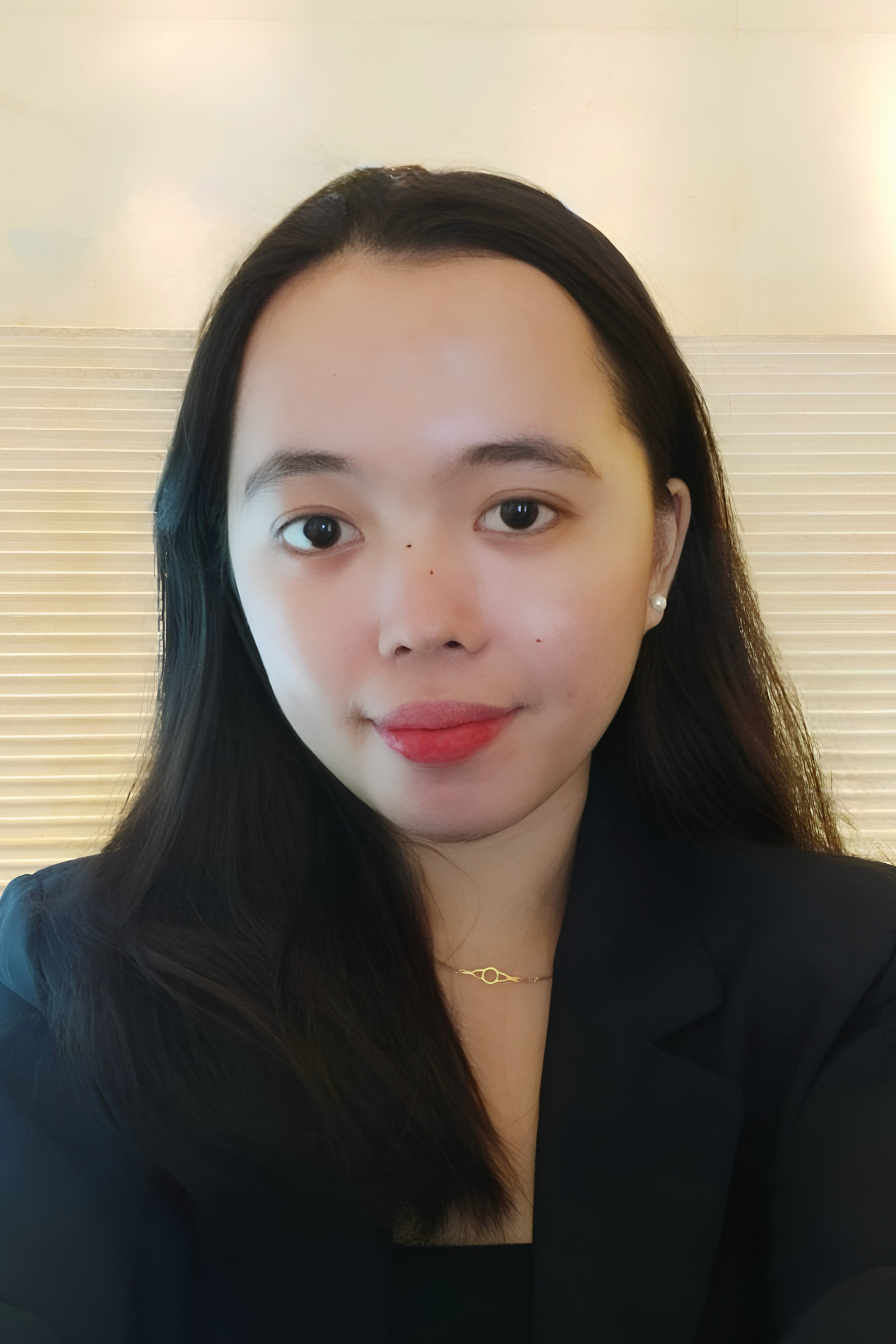 A woman with black hair taking a selfie. She is wearing pearl earrings and a delicate chain necklace, with a blurred background that includes horizontal lines.
