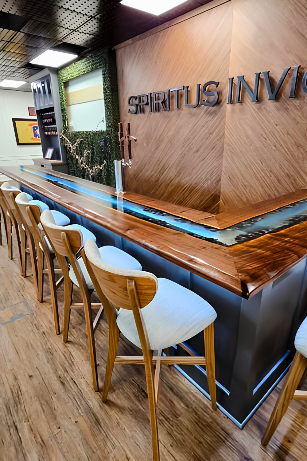 A bar interior with a wooden counter and white barstools. The wall behind features the words 'SPIRTUS INVI' in metallic letters. The decor includes greenery and artwork on the walls.