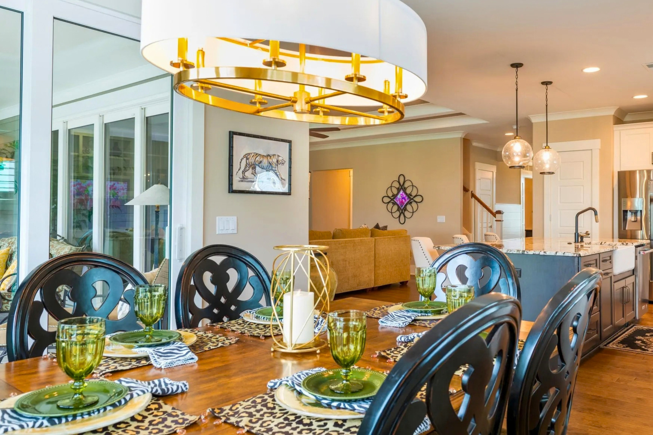 A dining area with a wooden table set with green glasses, leopard print placemats, and blue and white napkins, surrounded by black chairs, with a gold and white modern chandelier above. The background features a living room with beige walls, wall art, and a kitchen with white cabinets.