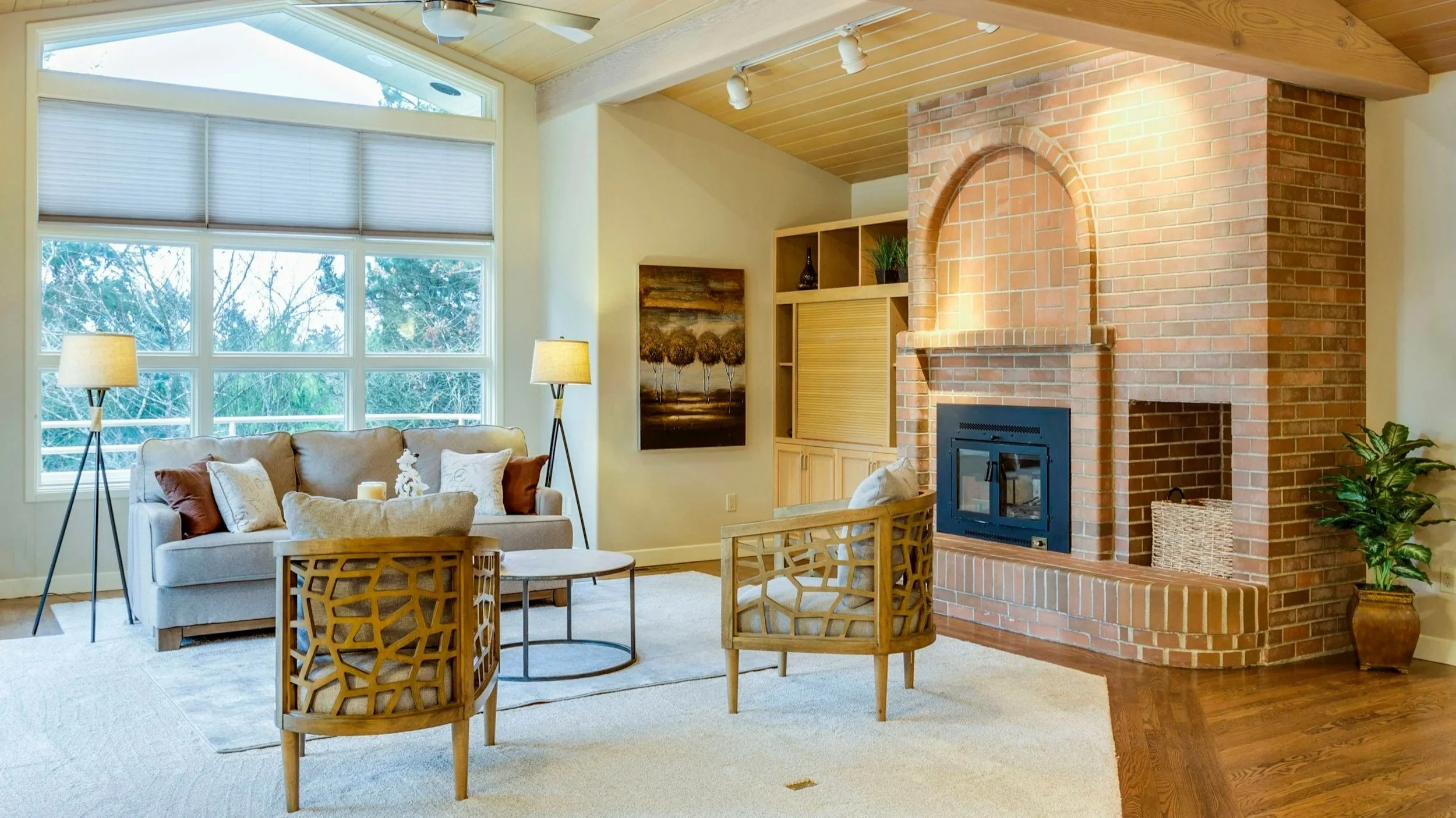 Living room with large windows, beige sofa with pillows, wooden and metal chairs, a brick fireplace, and lamps, with a ceiling fan and wood-paneled ceiling.