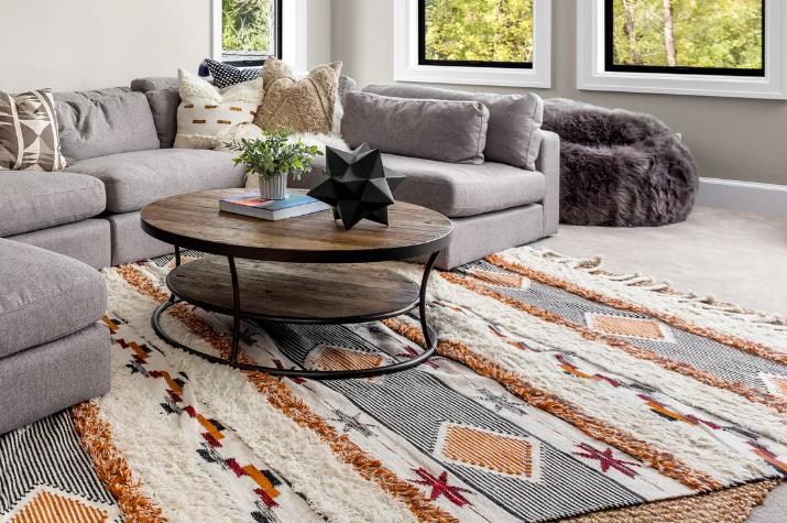 A stylish living room corner featuring a patterned, shaggy area rug with orange and grey accents layered over a jute rug, centered under a two-tiered wooden coffee table and a grey sectional sofa.