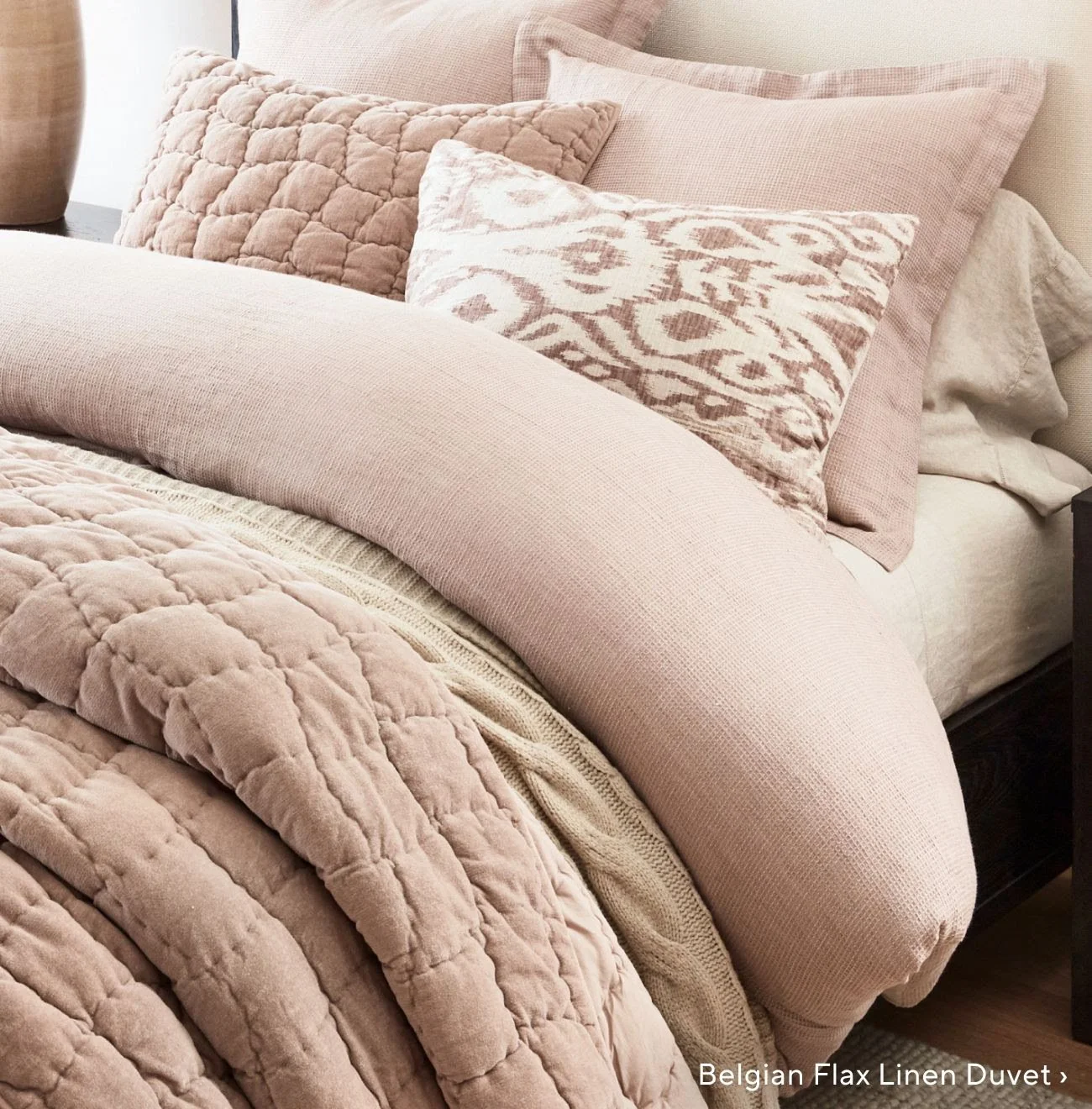 A close-up shot of a cozy bed styled with layers of dusty rose quilted and waffle-weave linens, a cream cable-knit blanket, and a textured geometric ikat accent pillow.