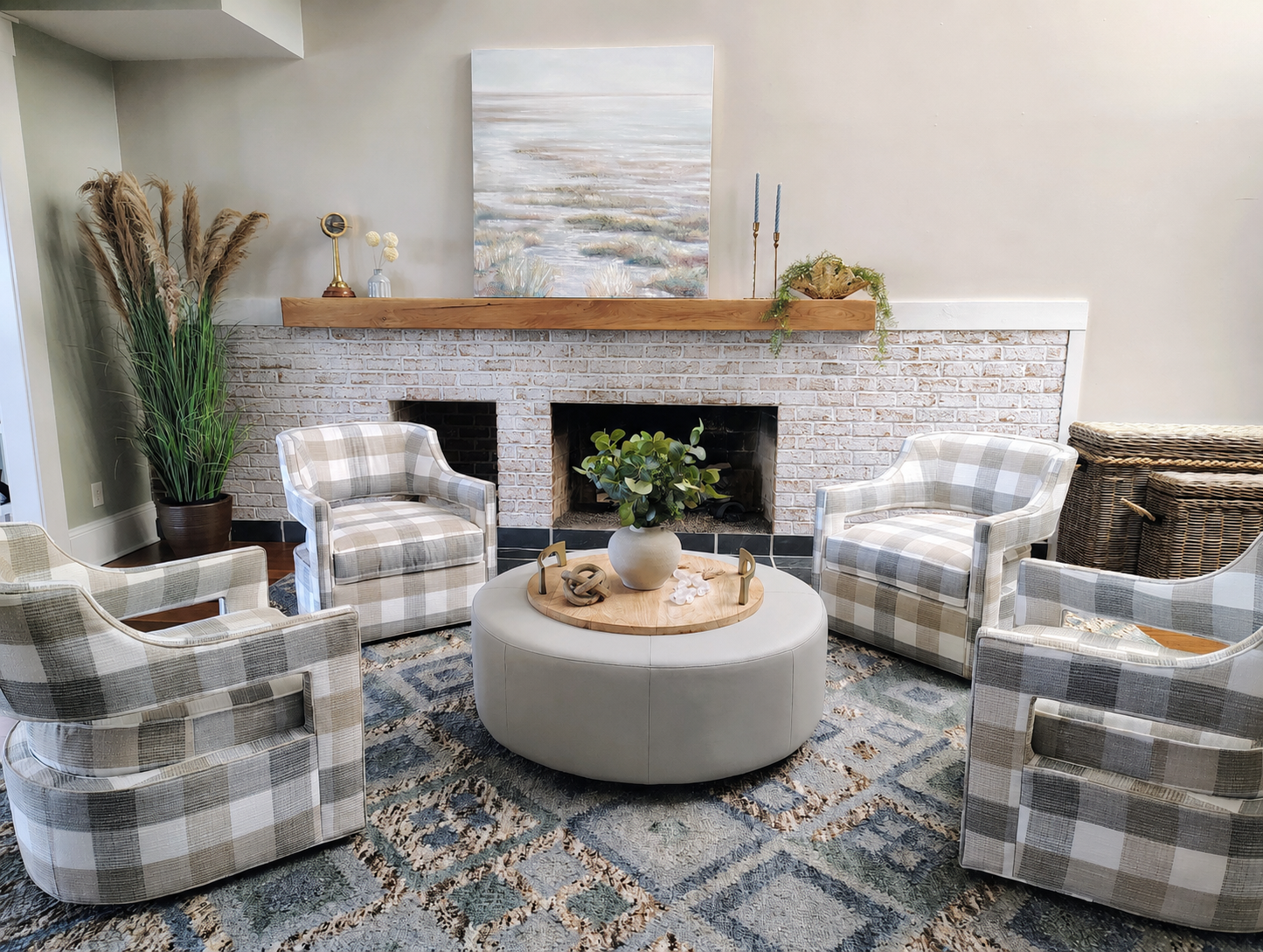 Living room with four plaid armchairs arranged around a round white ottoman with a plant on top, a brick fireplace with a wooden mantel, and decorative items including candles, a painting, and plants.
