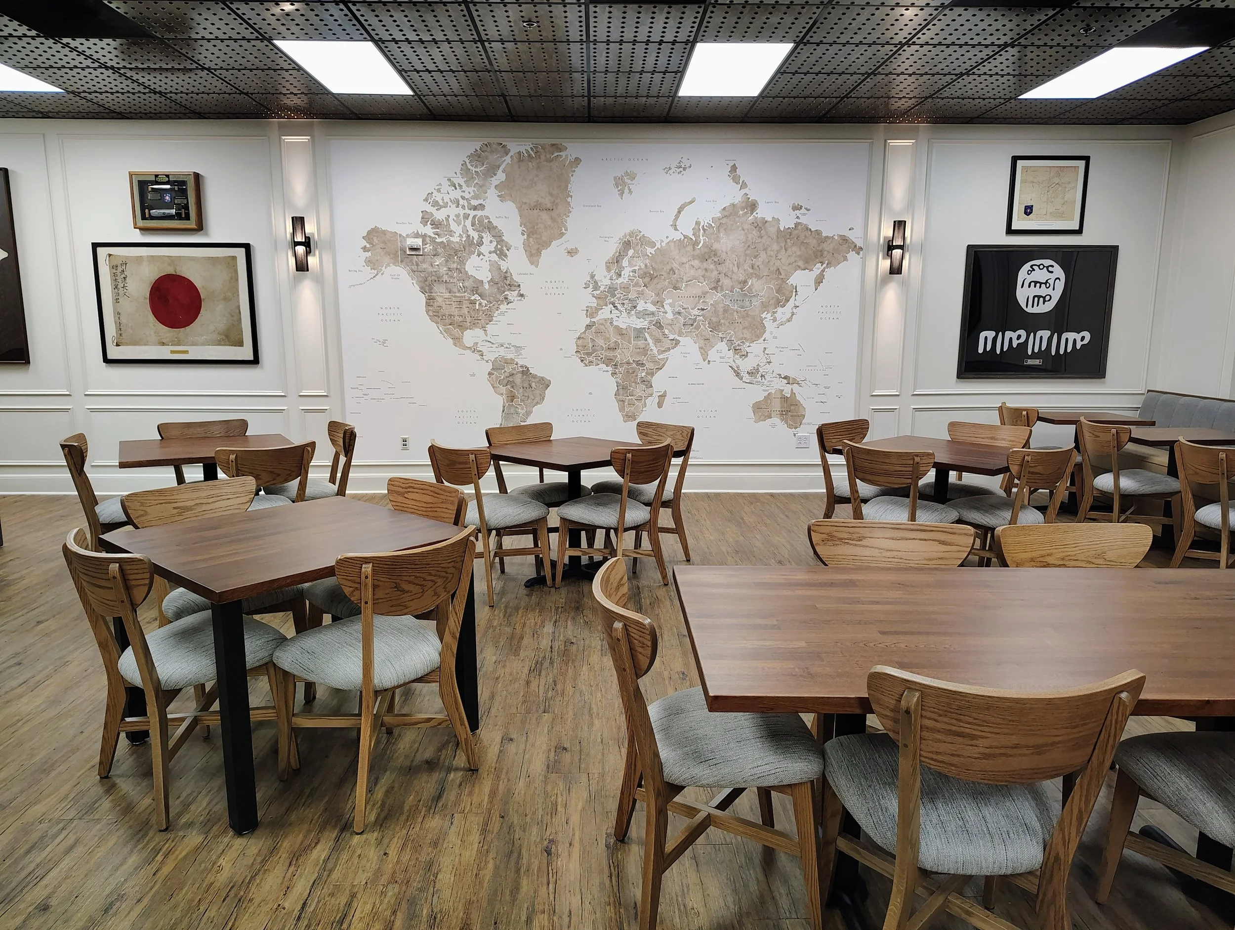 A room with wooden tables and chairs, a large world map on the wall, and framed artwork, including a Japanese flag and some documents. The ceiling has square lights and black perforated panels.