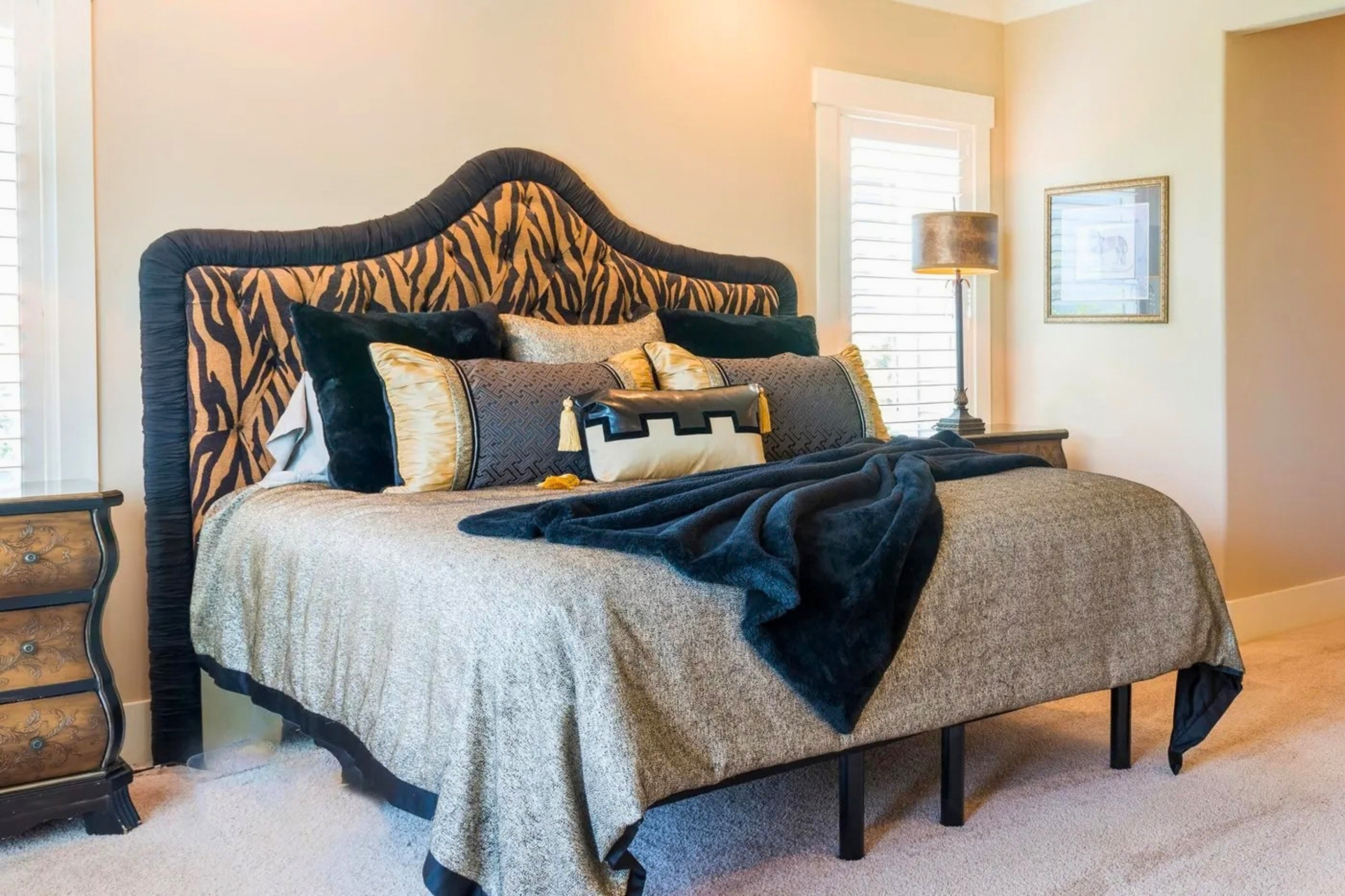 A bedroom with a large bed featuring a zebra-patterned headboard and various decorative pillows. A bedside table with a metallic lamp and a framed picture on the wall.