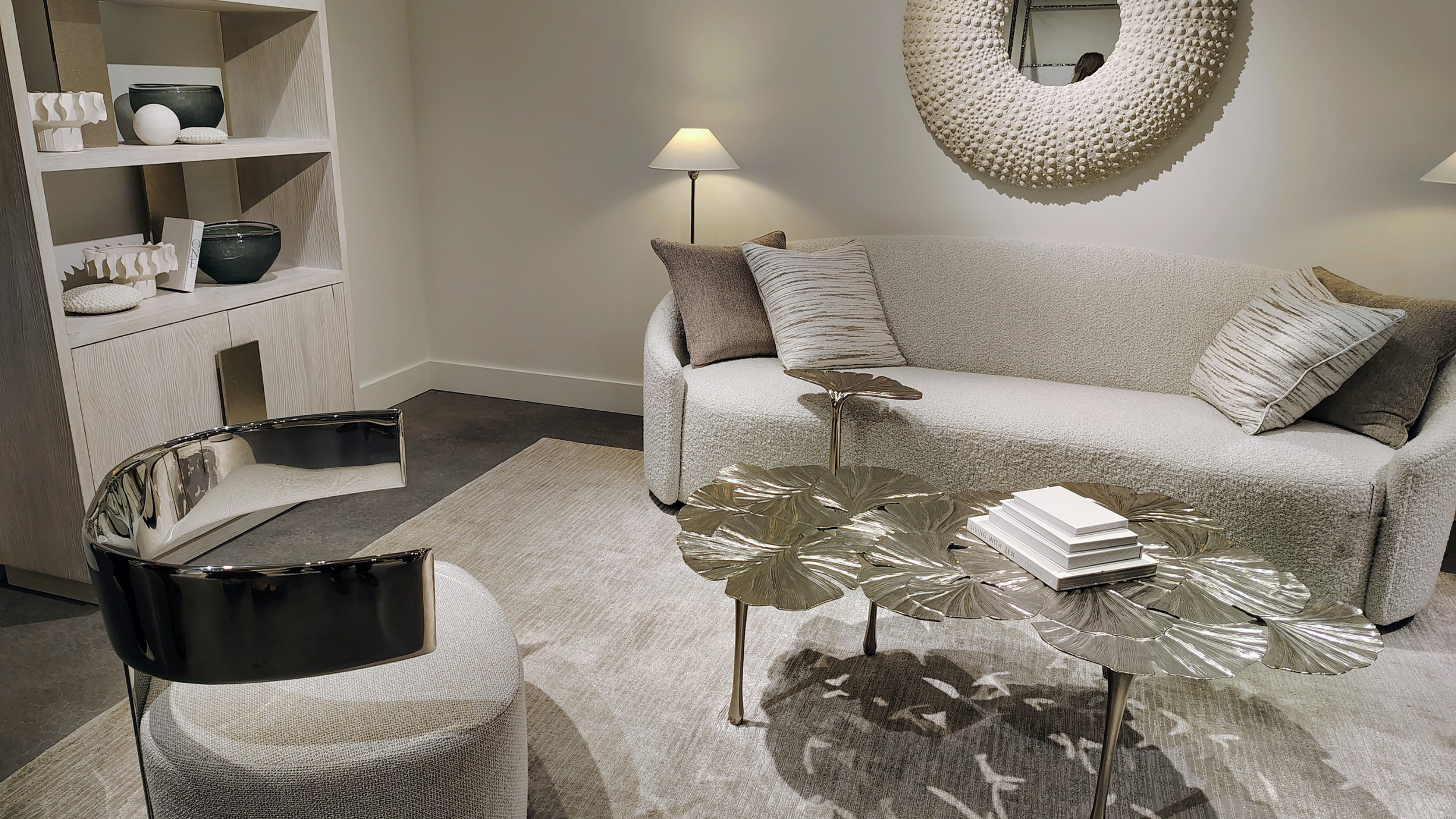 A modern living room with a beige sofa, decorative pillows, a round brass coffee table with a leaf pattern, a gold and black chair, a bookshelf with vases, a wall mirror with a textured frame, and a floor lamp, all on a neutral rug.