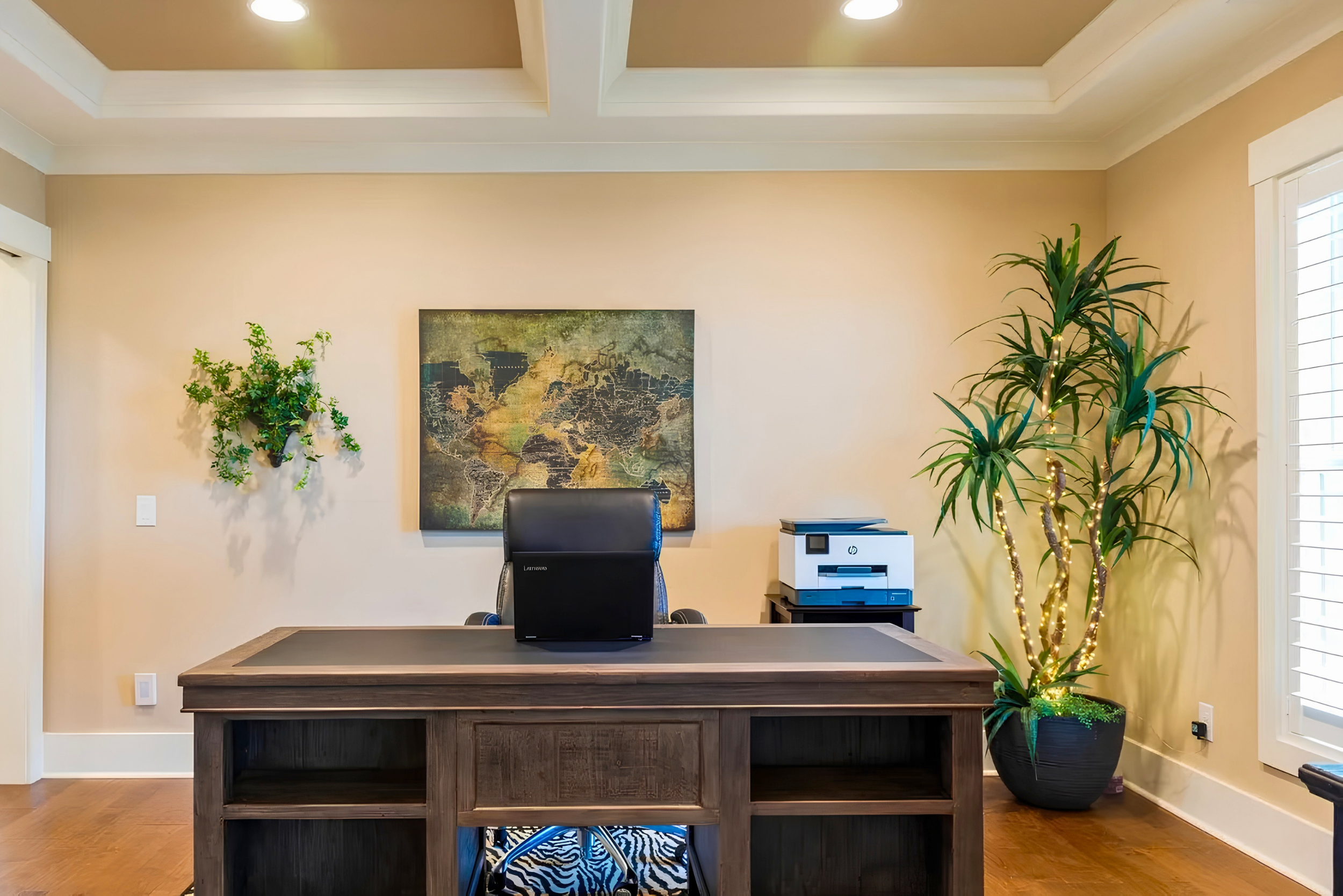 Home office with a dark wood desk, black office chair, artwork of a map on the beige wall, green plants on either side, and window with white blinds.