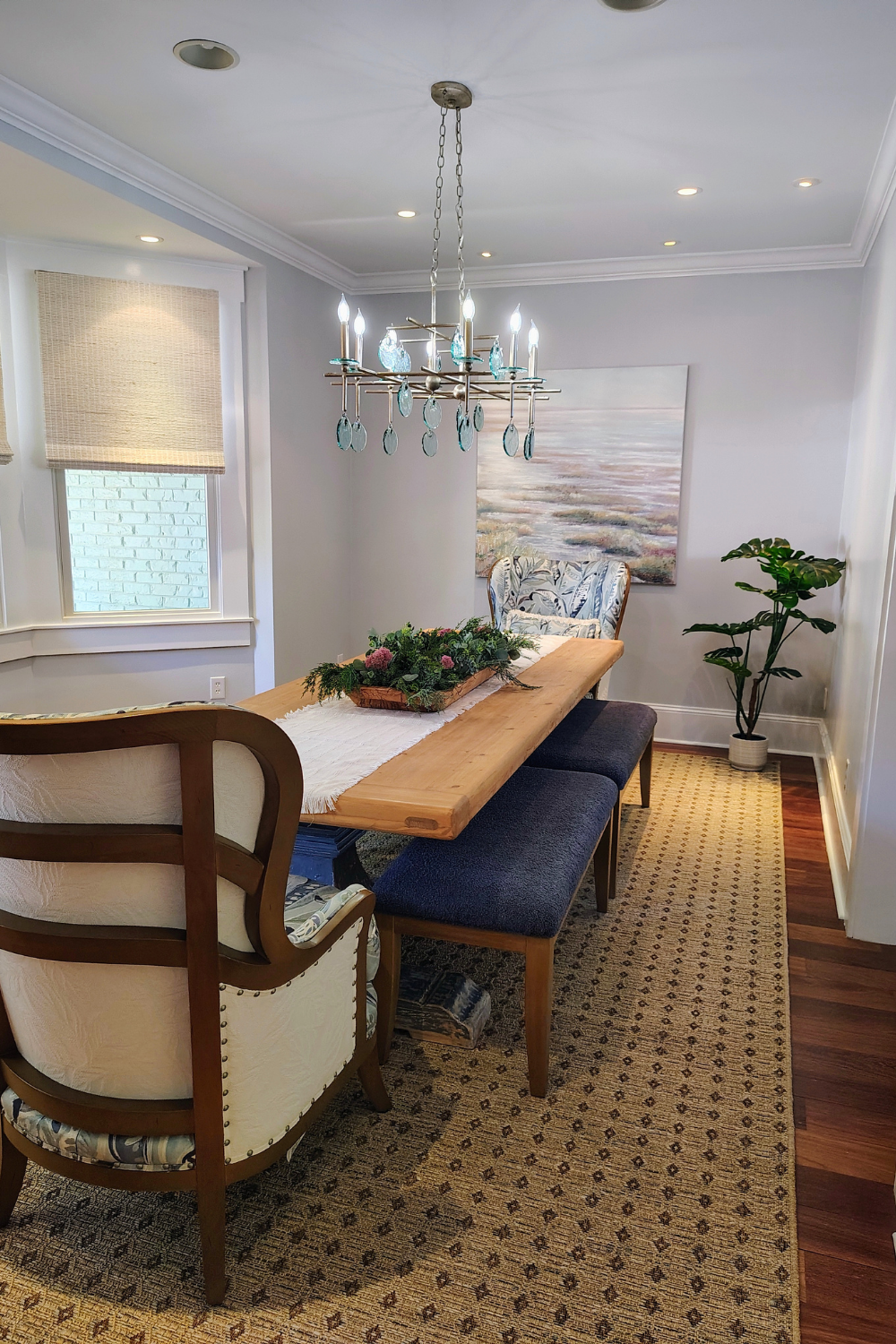 A dining room with a wooden table, a floral centerpiece, a patterned armchair, a large window with a blind, a potted plant, and a chandelier with teal glass ornaments.