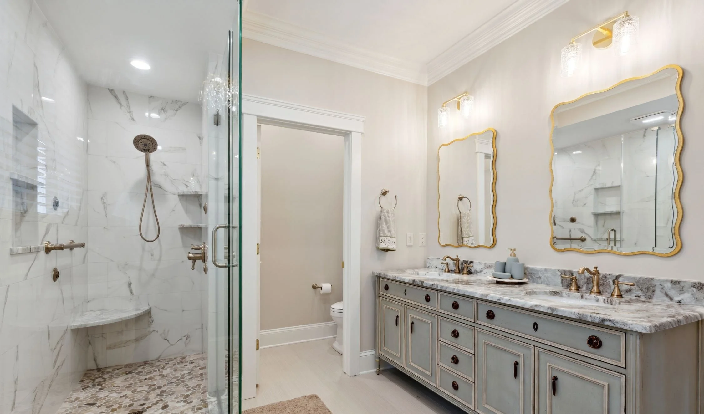 A bathroom with a marble vanity, gold fixtures, two mirrors with gold frames, a marble shower with glass door, and beige walls.