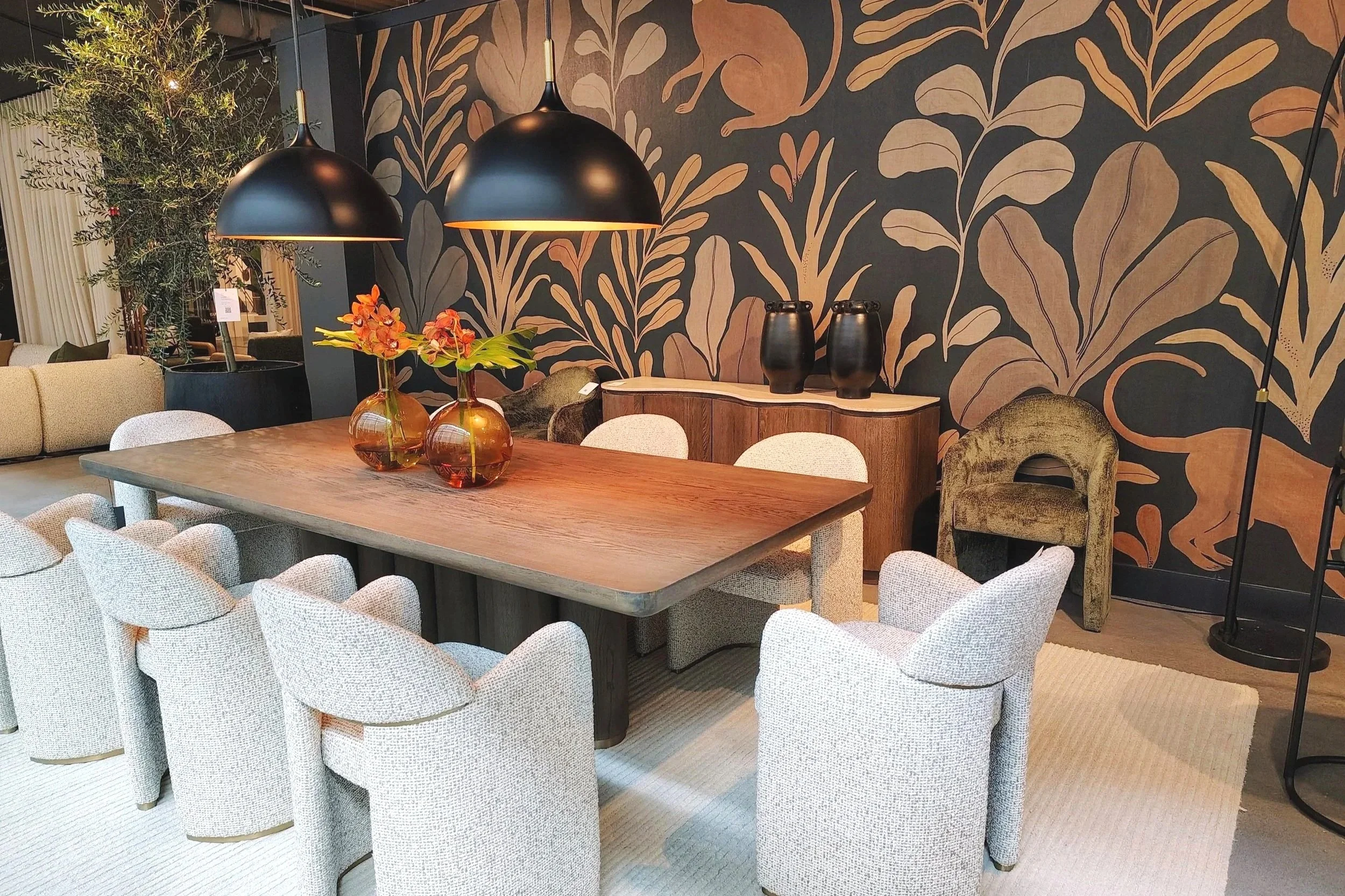 Interior of a modern dining room with a wooden table, surrounded by white upholstered chairs, a large black and gold pendant light, a botanical wallpaper mural, and decorative vases on a sideboard.