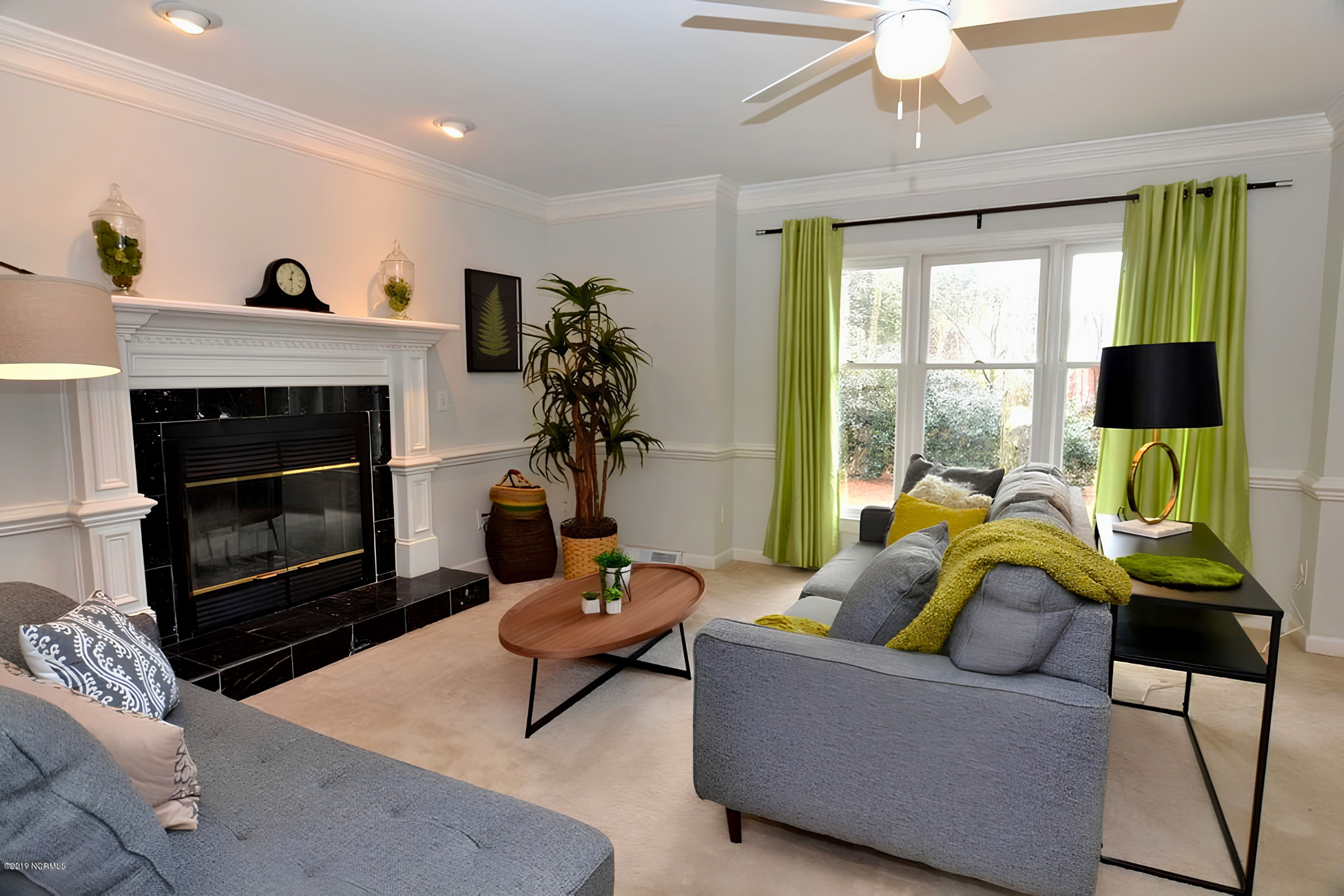 Living room with cream-colored walls, a fireplace with a black tile surround, a gray sofa with yellow and gray pillows, a black side table with a lamp and decor, a tall indoor plant, large windows with green curtains, and a ceiling fan with lights.
