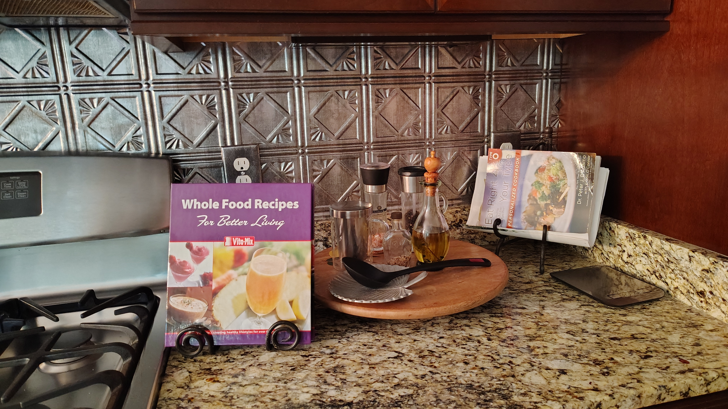 Kitchen countertop with spice jars, a cutting board, and a cookbook stand holding a cookbook titled 'Eat Right Your Type'. There is a purple brochure titled 'Whole Food Recipes For Better Living' and a glass dish with some oil or vinegar. The background features a decorative metallic tiled wall.