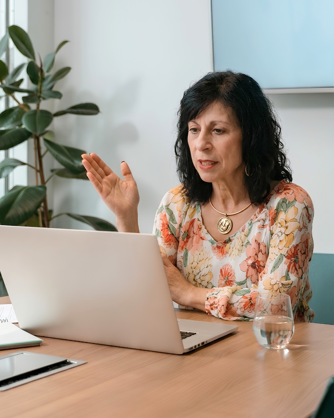 A woman with black curly hair wearing a floral blouse and gold jewelry is gesturing with her hand while seated at a wooden table with a laptop, a glass of water, and a tablet in front of her in a well-lit room with a potted plant and a blue wall.