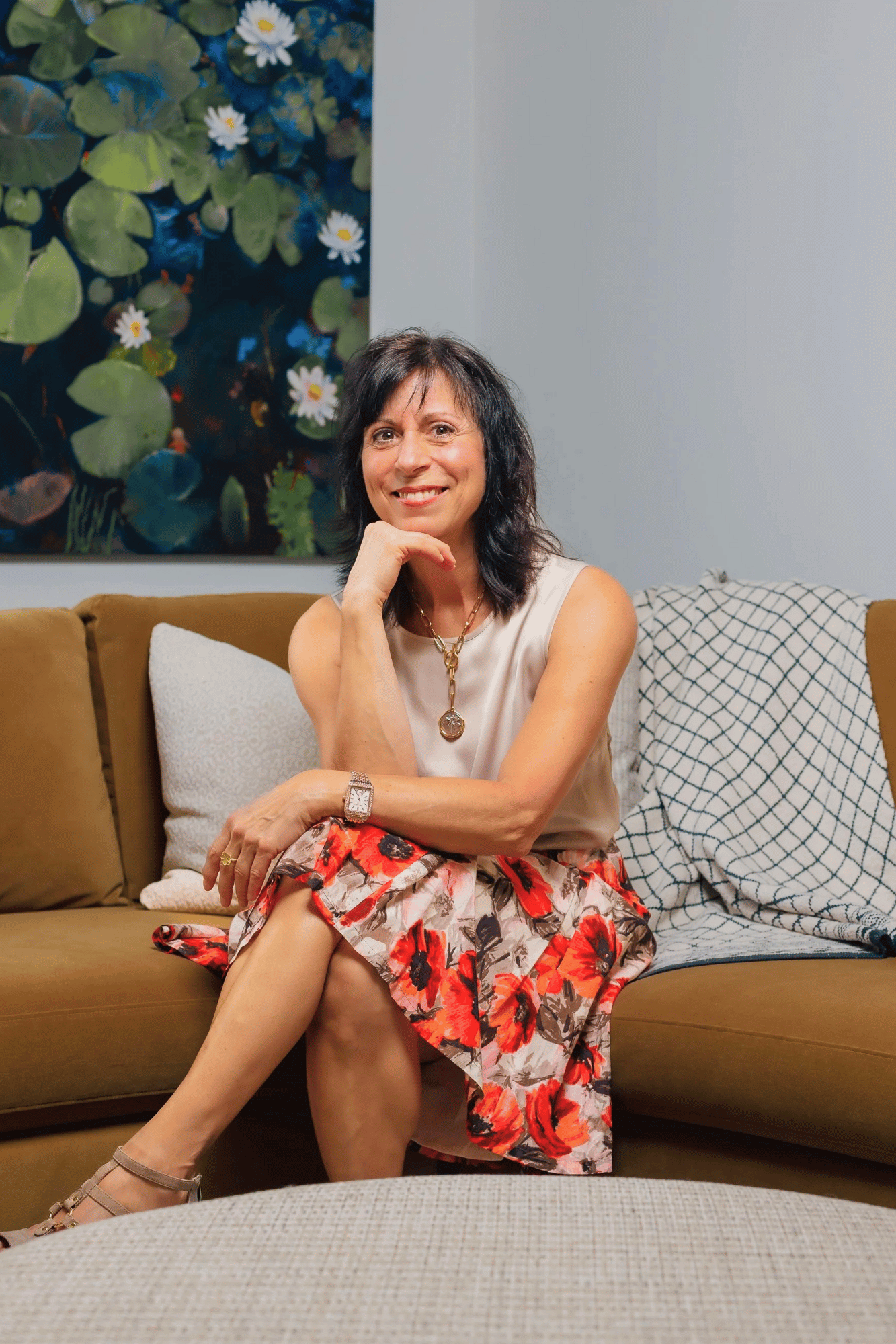 A woman with dark hair, wearing a beige sleeveless top, a colorful floral skirt, and jewelry, sitting on a mustard-colored sofa with her legs crossed. She's smiling with her chin resting on her hand. Behind her is a large painting of water lilies and a white wall, with patterned pillows on the sofa.