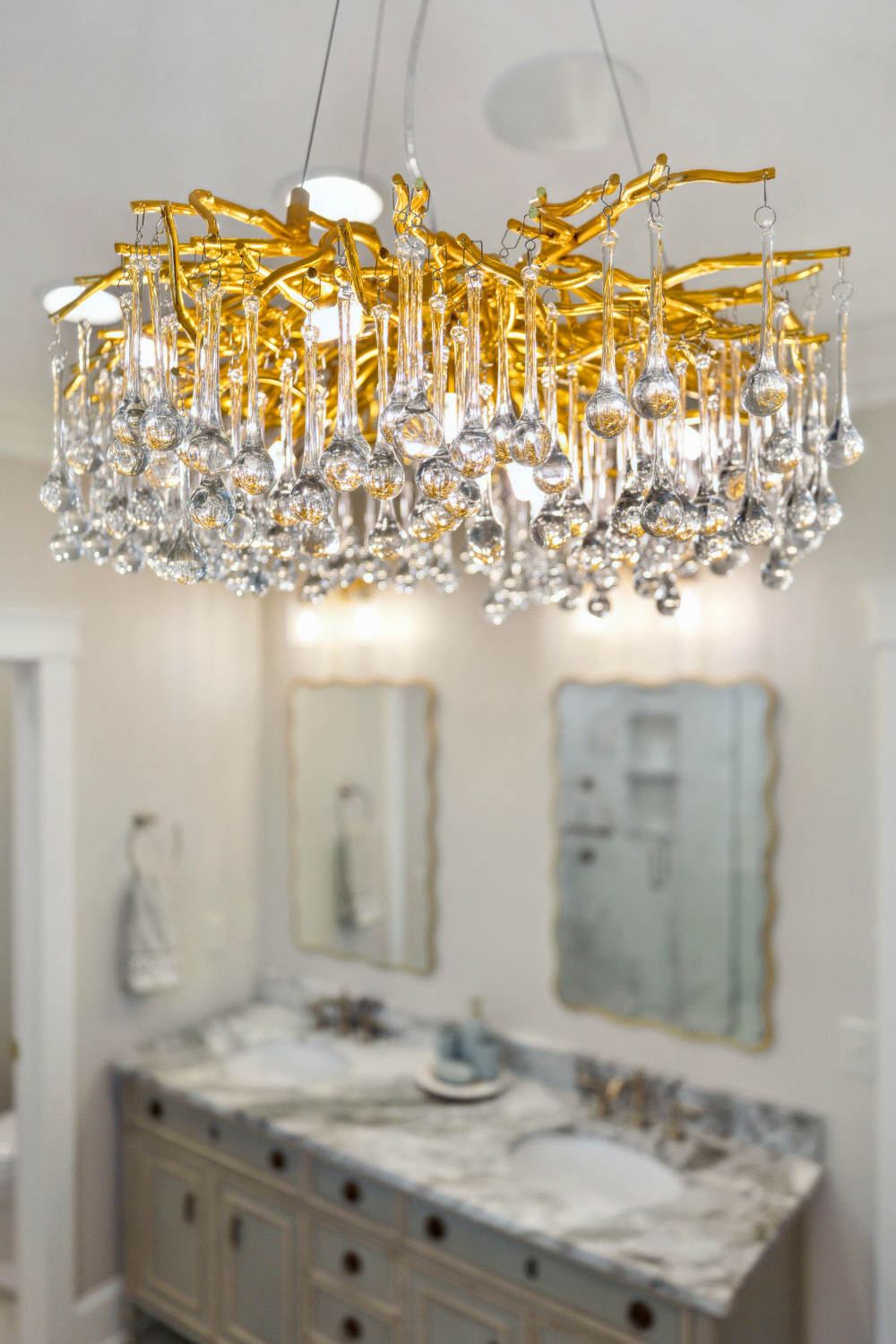A luxurious chandelier with gold accents and hanging crystals in a bathroom with double mirrors and a marble countertop.