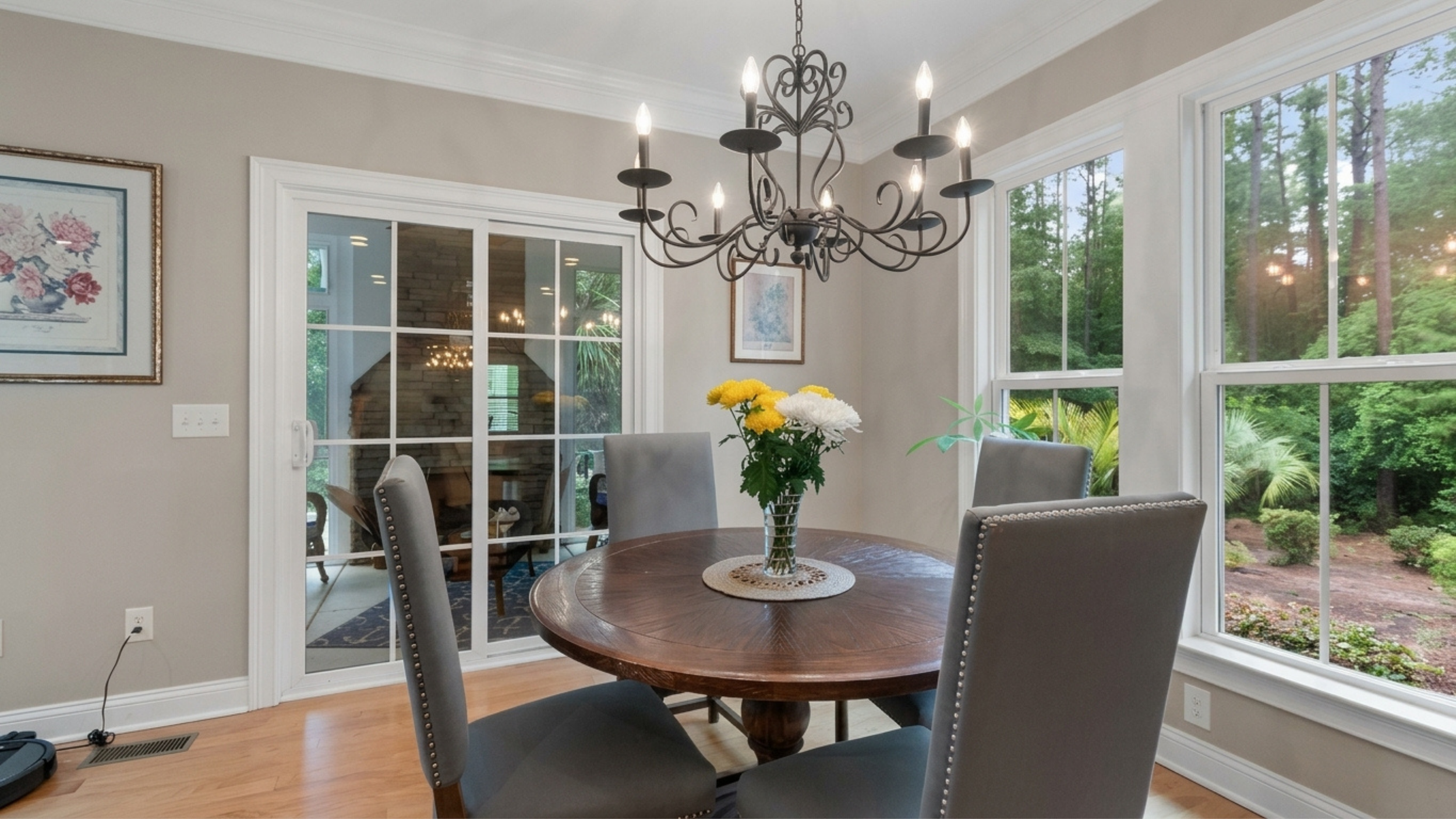 Dining room with a wooden table, gray upholstered chairs, a black chandelier, and large windows showing a green outdoor landscape.