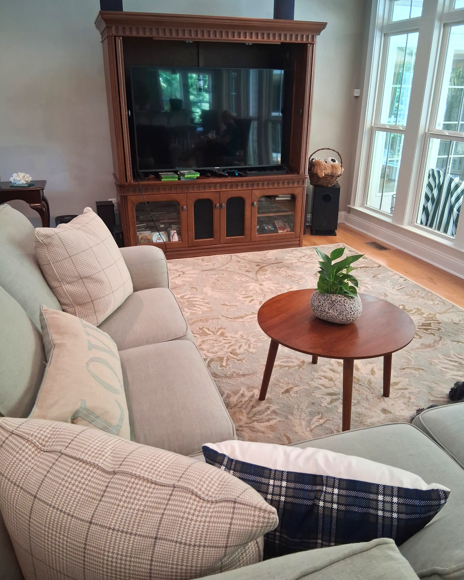 Living room scene featuring beige and plaid-patterned couches, a round wooden coffee table with a potted plant, a large flat-screen TV on a wooden stand, a basket with toys, and large windows letting in natural light.