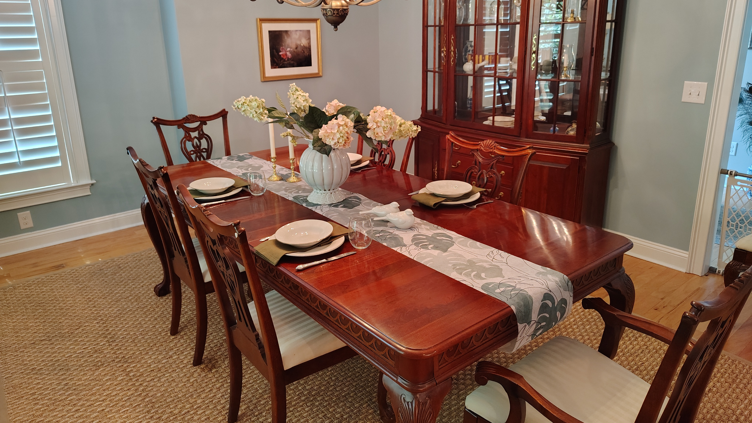 A dining room with a rectangular wooden table set with white plates, glassware, silverware, cloth napkins, and a floral table runner. In the center, there is a large vase with pink and white hydrangeas and two lit candles in candlesticks. The room features a wooden china cabinet filled with dishes and decorative items. There are six matching wooden chairs with beige upholstered seats around the table. The walls are painted light blue, and there is a window with white shutters on the left. A framed picture hangs on the wall, and a brass chandelier hangs above the table. The floor is hardwood with a woven beige rug underneath the table.