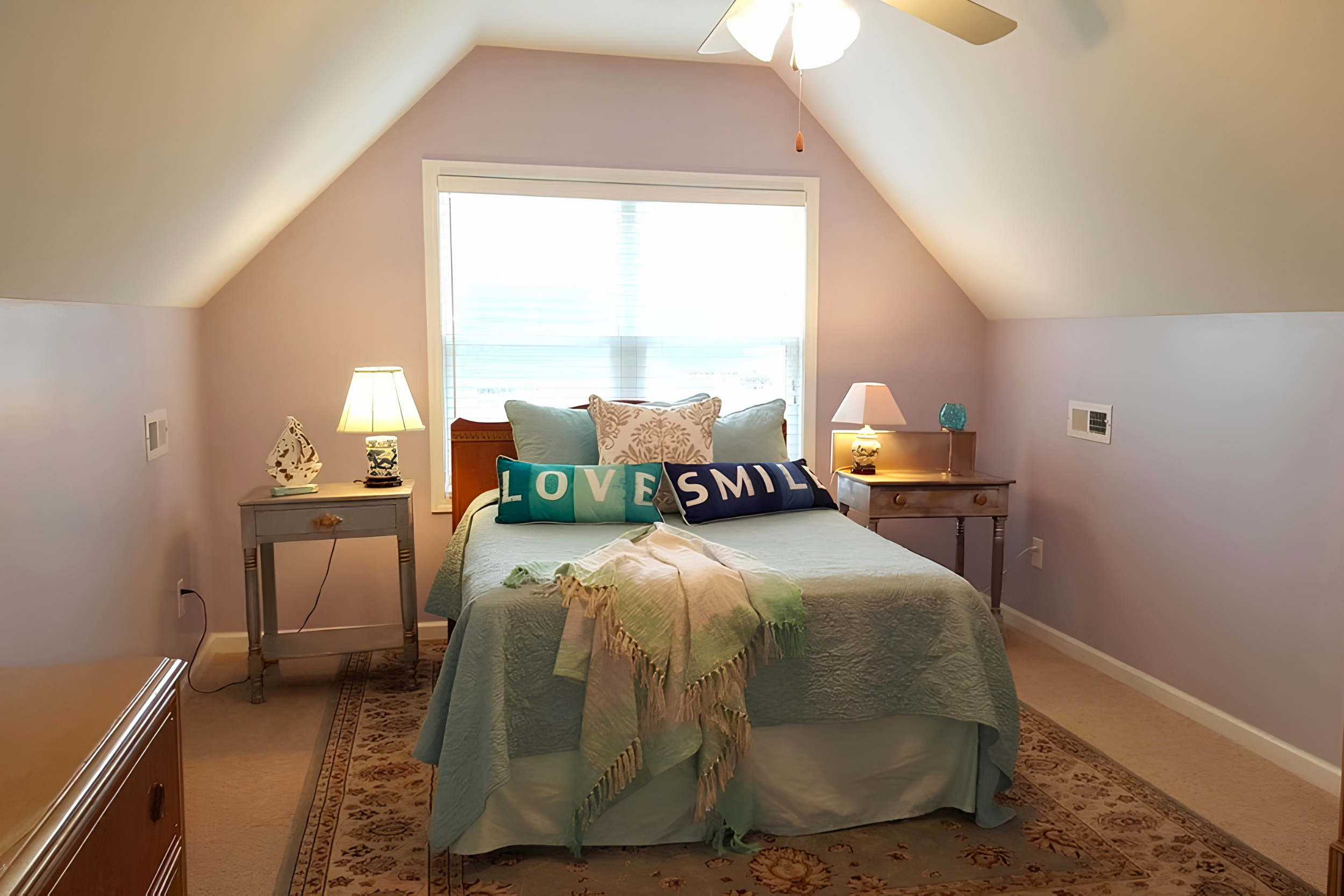 A bedroom with a sloped ceiling, a large window, and a bed with decorative pillows that say 'LOVE' and 'SMILE'. Two nightstands with lamps are on either side of the bed. A small table with a decorative item and a lamp is on the left, and a small table with a decorative item and a lamp is on the right. A ceiling fan is above, and an area rug is on the carpeted floor.