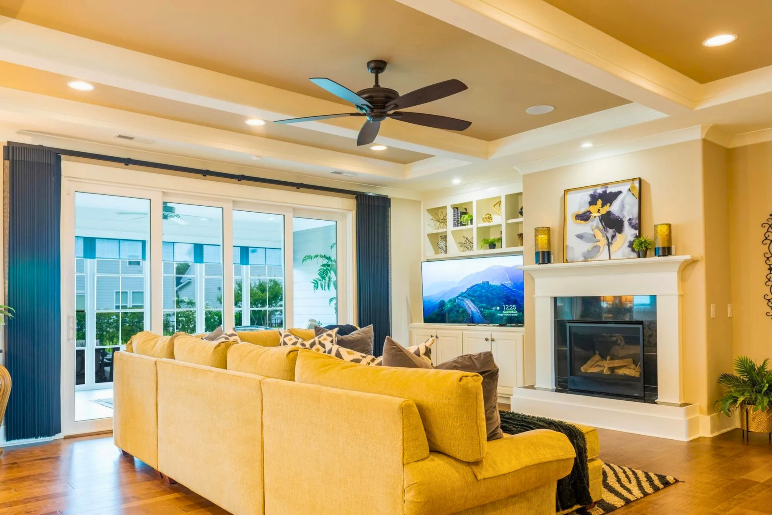 Living room with a yellow sofa, a fireplace, a mounted flat-screen TV, and large windows with sliding glass doors leading to a patio.